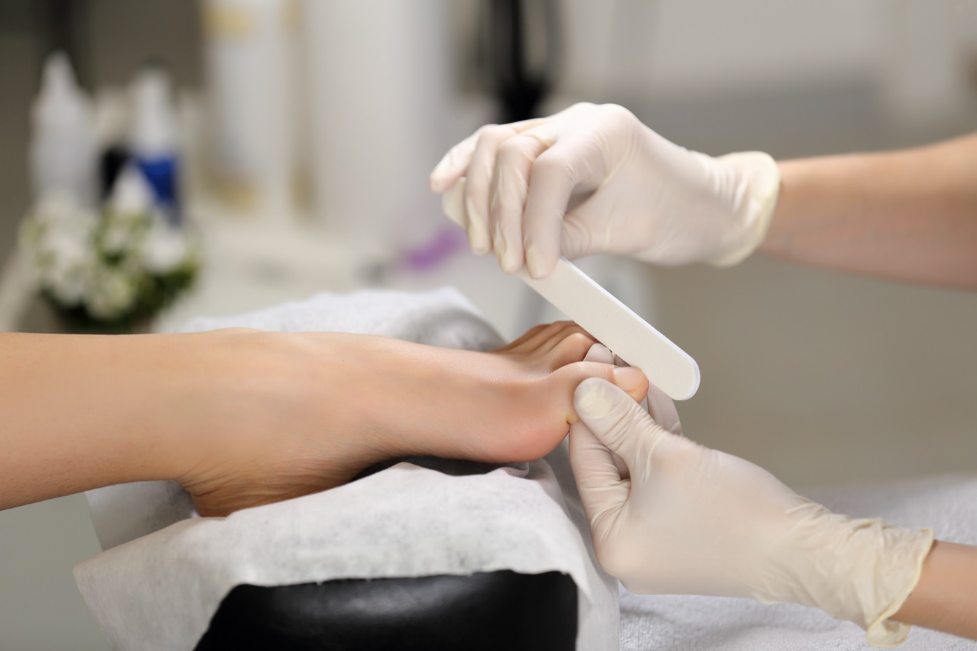 A nail technician wearing gloves files a client's toenail during a pedicure